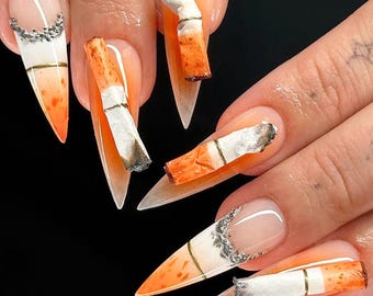 May include: Close-up of several long, pointed fingernails with orange and white nail polish. Each nail is decorated with a miniature cigarette, some with the filter end visible.