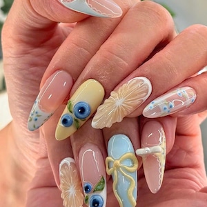 Puede incluir: Una mano que muestra uñas largas en forma de almendra con diseños detallados. Las uñas están pintadas en rosa, amarillo y azul, con adornos 3D como lazos, arándanos y rodajas de cítricos. Algunas uñas tienen delicados diseños florales.