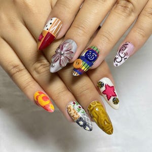 May include: A set of long, almond-shaped fingernails with unique, colourful designs. Each nail features a different pattern, including patchwork, floral, and button embellishments. Colours include red, orange, yellow, white, and various shades of grey and brown.