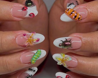 May include: Close-up of a hand with several oval-shaped nails painted with a white tip and decorated with various 3D insect and flower designs. The designs include a ladybug, a caterpillar, a flower, a beetle, and an ant.