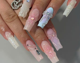 May include: Close-up of long, nude-colored acrylic nails with 3D floral and bow designs. The nails are decorated with rhinestones and pearls.
