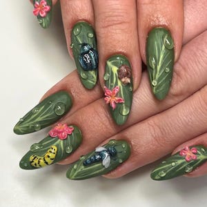 Puede incluir: Uñas verdes en forma de almendra con diseños de hojas y gotas de agua. Algunas uñas presentan insectos en 3D y flores rosas. Las uñas tienen un acabado brillante, creando una estética inspirada en la naturaleza.
