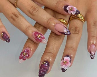 May include: Close-up of hands with burgundy and nude almond-shaped nails decorated with 3D flowers. The flowers are pink, white, and red with gold accents.