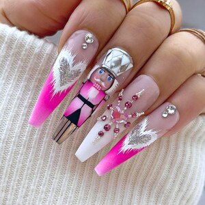 May include: Pink and white ombre long false nails with a nutcracker design on one nail and a snowflake design with rhinestones on another nail.
