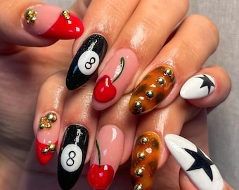 May include: A hand displaying a set of long, decorated fingernails. The nails feature various designs, including a red and pink color block, a black eight ball, a cherry, a leopard print, and a white nail with a black star. Some nails have gold embellishments.