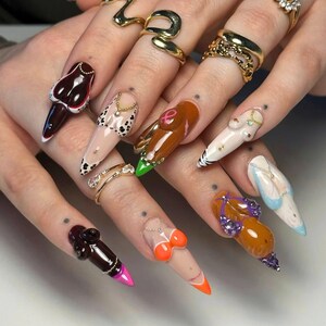 May include: A close-up of a hand with long, sculpted fingernails. Each nail features a unique design, including miniature sculptures of body parts, animal prints, and abstract art. The nails are adorned with various colours and textures, and some have small jewellery accents.