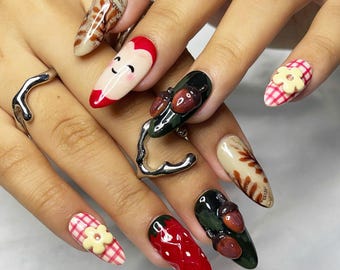Könnte beinhalten: Nahaufnahme einer Hand mit langen, verzierten Fingernägeln. Die Nägel weisen verschiedene Designs auf, darunter eine rote Erdbeere, ein lächelndes Gesicht, Eicheln und eine Blume. Die Nägel sind in verschiedenen Farben bemalt, darunter Rot, Grün, Beige und Weiß. Ein silberner Ring ist ebenfalls sichtbar.