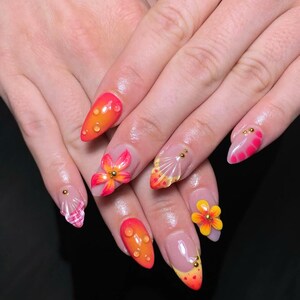 Könnte beinhalten: Ein Set langer, mandelförmiger Acrylnägel mit verschiedenen Designs. Einige Nägel zeigen 3D-Blumen, während andere Wassertropfeneffekte oder Muschelmuster aufweisen. Die Farbpalette umfasst Orange, Gelb, Rosa und Transparent.