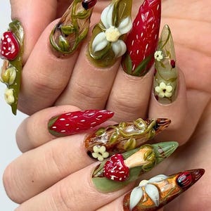May include: A set of long, pointed acrylic nails with intricate designs. The nails feature a variety of elements, including strawberries, flowers, ladybugs, and vines, in shades of red, green, white, and brown. The designs are detailed and artistic.