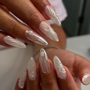 May include: Close-up of a hand with long, pointed nails. Each nail is painted with a pearly white polish and decorated with a 3D design resembling a seashell. Some nails have small pearl accents.