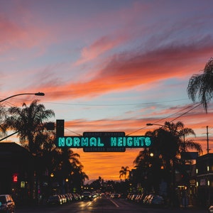 Puede incluir: Un letrero de ne&oacute;n con las palabras "NORMAL HEIGHTS BUSINESS DISTRICT" contra un cielo de puesta de sol vibrante. Palmeras bordean la calle en silueta.