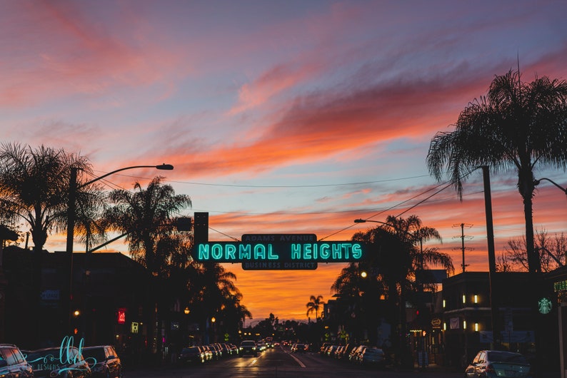 Puede incluir: Un letrero de ne&oacute;n con las palabras "NORMAL HEIGHTS BUSINESS DISTRICT" contra un cielo de puesta de sol vibrante. Palmeras bordean la calle en silueta.