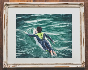 Può includere: Una persona con una muta da surf nera e un giubbotto giallo fa surf su una tavola da surf bianca in acqua blu.