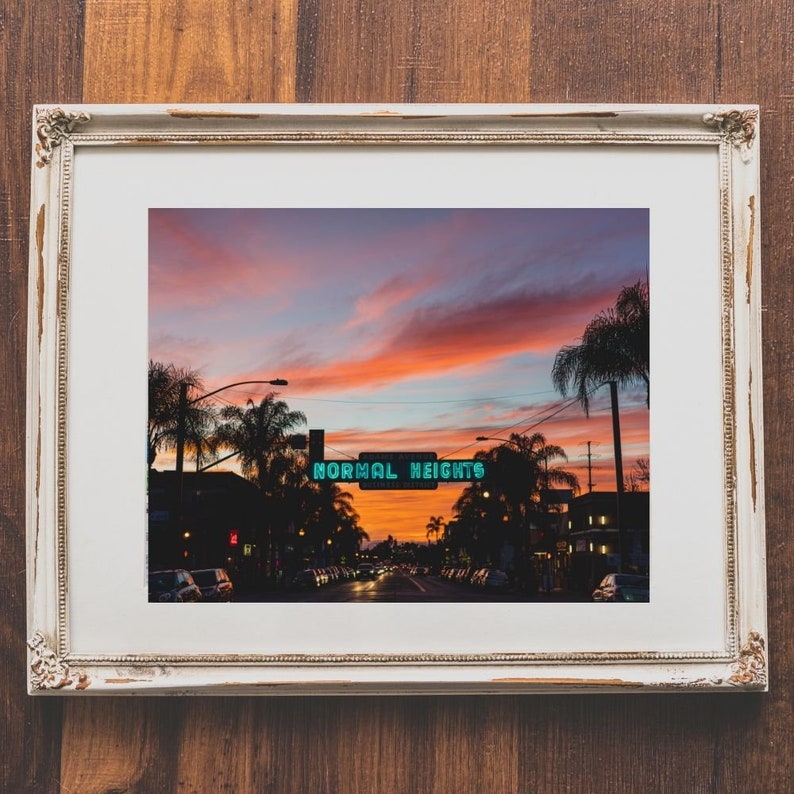 Puede incluir: Una fotograf&iacute;a enmarcada de una escena callejera al atardecer. La calle est&aacute; bordeada de palmeras y el cielo es de un naranja y rosa vibrantes. Un letrero de ne&oacute;n dice "NORMAL HEIGHTS".