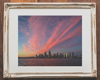 May include: A framed photograph of a city skyline at sunset. The sky is a vibrant pink and orange, with clouds stretching across the horizon. The city buildings are silhouetted against the sky.