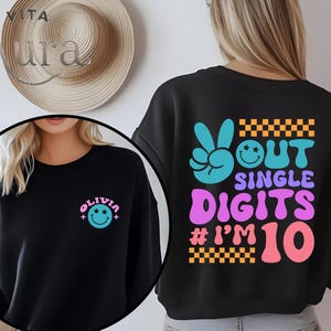 May include: Black sweatshirt with a colorful retro design. The front features a name and a smiley face. The back has a peace sign, smiley face, and the words "OUT SINGLE DIGITS # I'M 10" in a groovy font.