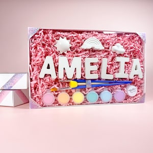 May include: A craft kit with the name "AMELIA" in white block letters, along with sun, rainbow, and cloud shapes. The kit includes paintbrushes, paint pots in various colors, and is packaged in a clear box with pink shredded paper.