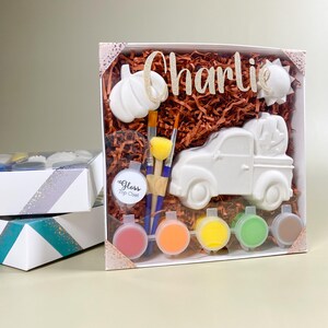 May include: A white ceramic paint-your-own kit with a truck carrying pumpkins, a pumpkin, and a sun. The kit includes paintbrushes, a sponge, and paint pots in various colors. The name "Charlie" is written in glittery gold.