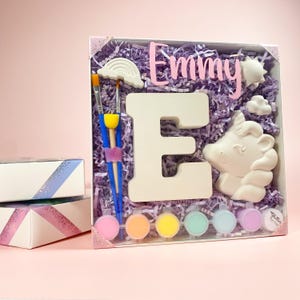 May include: A craft kit with a white ceramic letter E, a unicorn figurine, paint pots, paintbrushes, and a rainbow. The kit is packaged in a clear plastic box with purple tissue paper and the name "Emmy" written in pink letters.