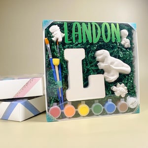 May include: A paint your own ceramic kit featuring the letter L, a dinosaur, and a small dinosaur figurine. The kit includes six paint pots, two paintbrushes, and green shredded paper. The name Landon is written in green letters on the box.