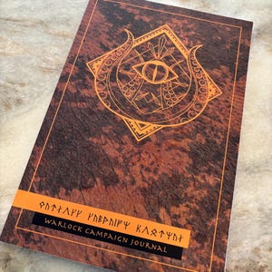May include: A brown and orange journal with a gold eye and tentacles design. The text "Warlock Campaign Journal" is printed on the front.