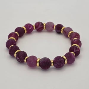 Può includere: Un braccialetto di perline con grandi perline rotonde viola con una superficie strutturata. Le perline sono separate da piccoli distanziatori metallici color oro. Il braccialetto è di colore viola scuro.
