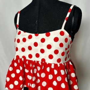 May include: A sleeveless peplum top featuring a cream-coloured bodice with red polka dots and a red peplum hem with white polka dots. The garment has thin shoulder straps and a straight neckline, displayed on a black mannequin torso.