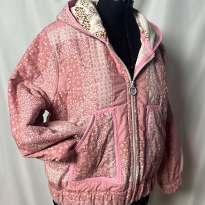 May include: A quilted pink jacket with a hood, featuring a patchwork design of floral patterns. The jacket has a full zipper, two front pockets, and a contrasting floral print lining in the hood. The jacket is displayed on a black mannequin.