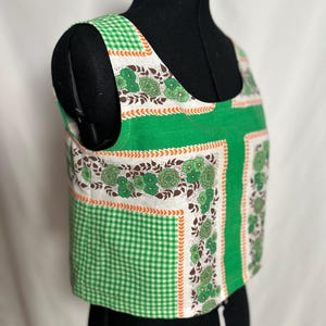 May include: A sleeveless crop top featuring a vibrant green and white gingham pattern combined with panels of white fabric decorated with a brown and green floral print and an orange leaf-like border detail, finished with a wide scoop neckline.