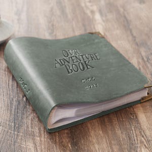 以下が含まれることがあります： 「Our Adventure Book」と「Vol. 1」のエンボス加工が施された、緑色の革製アドベンチャーブック。真鍮製のコーナープロテクターが付いており、2025年も表紙にエンボス加工されています。本は開かれており、白いページが見えています。