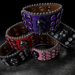 Op de afbeelding: Een verzameling leren armbanden in verschillende kleuren, waaronder paars, rood, roze en zwart. Elke armband heeft een schedelontwerp en is versierd met strass-steentjes en metalen studs. De armbanden hebben een punkrock-esthetiek.
