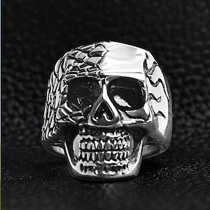 May include: A silver skull ring with detailed facial features, including eye sockets, a nose cavity, and teeth. The ring has a textured design on the sides and top, creating a gothic or biker aesthetic.