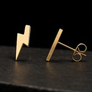 May include: A pair of gold-colored stud earrings. One earring is a lightning bolt shape, and the other is a straight bar. The earrings are set against a dark background, highlighting their metallic sheen. These earrings are a stylish accessory.