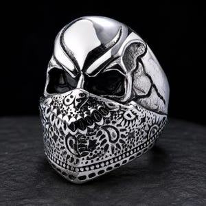 May include: A silver-toned metal ring shaped like a human skull. The skull features deep, dark eye sockets and is wearing a bandana across the lower face. The bandana is decorated with a detailed paisley and star pattern in a relief texture.