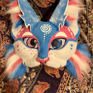 May include: A handmade furry mask in blue, pink, and white. The mask has large eyes, a pink nose, and fluffy fur around the ears and cheeks. The design includes painted details and a spiral on the forehead, creating a whimsical look.