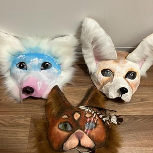 May include: Three handmade animal masks. The top left mask is white and blue, resembling a fantasy creature. The top right mask is tan and white with large ears. The bottom mask is brown with green eyes and feathers.