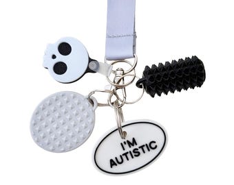 May include: A white lanyard with a variety of charms. Charms include a white skull, a textured oval, a black spiky cylinder, and an oval with the text "I'M AUTISTIC" in black. The charms are attached to a silver key ring.