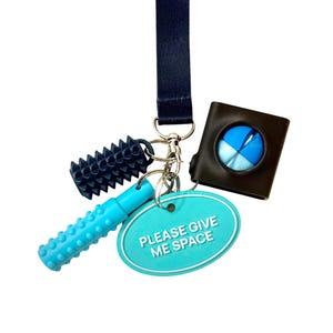 Op de afbeelding: Een verzameling sensorisch speelgoed en accessoires die aan een marineblauwe lanyard hangen. Inclusief een zwarte getextureerde roller, een lichtblauwe getextureerde buis, een bruin vierkant met een blauw-witte bal en een turquoise ovale tag met de tekst "PLEASE GIVE ME SPACE."