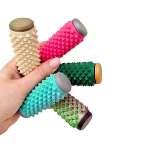 May include: Five colourful massage rollers are held in a hand. The cylindrical rollers have a textured, spiky surface. Colours include beige, pink, green, teal, and a green and purple gradient. Each roller has a different coloured end cap.