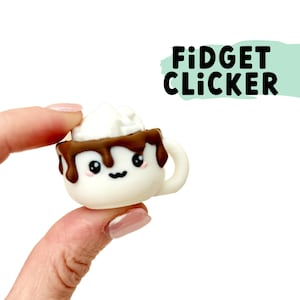 Puede incluir: Una taza de cerámica blanca con un chorrito de chocolate marrón y una crema batida blanca en la parte superior. La taza tiene una linda carita de dibujos animados con ojos y una sonrisa. La taza está etiquetada como "Fidget Clicker".