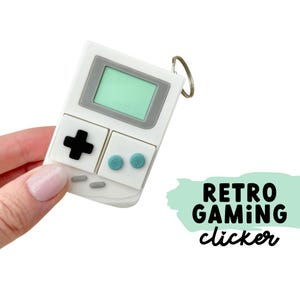 May include: White retro gaming clicker keychain with a green screen, grey border, black cross button, and teal buttons. The keychain has a silver ring and the words "Retro Gaming clicker" in black.