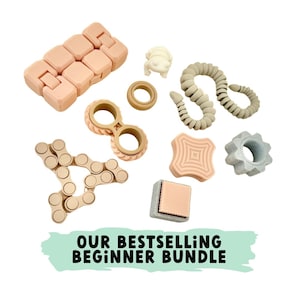 Puede incluir: Una colección de juguetes antiestrés de diferentes formas y colores, que incluyen un cubo rosa y beige, un anillo beige y dorado, una cadena beige y dorada, una estrella beige y dorada, un círculo beige y dorado, un cuadrado beige y dorado, una serpiente beige y dorada, una flor beige y dorada, un corazón beige y dorado y una mariposa beige y dorada. Los juguetes están dispuestos sobre un fondo blanco. Nuestro paquete de principiante más vendido.