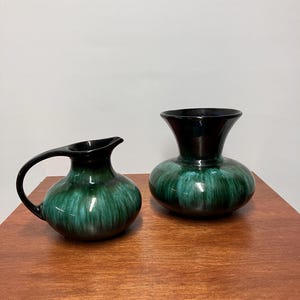 May include: A ceramic jug and a matching vase, both featuring a rounded body with a dark black rim that transitions into a vibrant, streaked emerald green glaze. The jug includes a curved handle and a spout, while the vase has a wide opening.