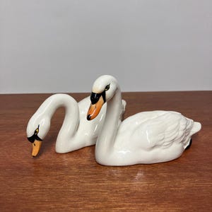 May include: Two white ceramic swan figurines with glossy finishes. One swan is positioned with its neck curved downwards, while the other swan stands upright with its neck arched. Both feature orange beaks with black accents and detailed eyes.