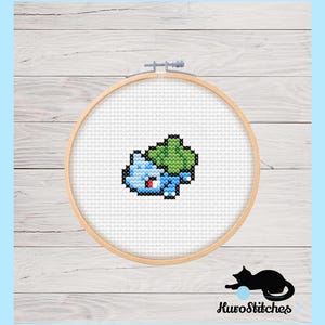 Op de afbeelding: Een kruissteekpatroon van een blauw en groen Pokemon-personage op een witte achtergrond, ingelijst in een houten hoepel.