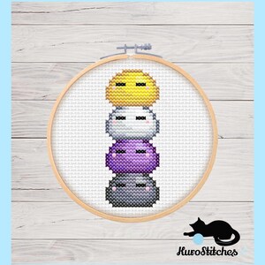 May include: A cross-stitch embroidery hoop showing a stack of four colourful, round characters. The characters are yellow, white, purple, and grey, each with closed eyes. The wooden hoop holds the fabric, set against a light blue background.