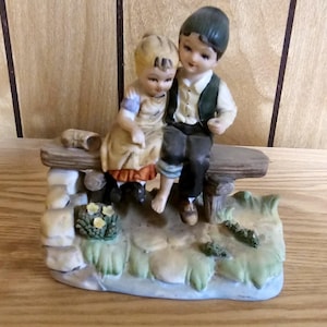 May include: A porcelain figurine depicting a boy and girl sitting on a bench. The boy wears a green vest and dark pants, while the girl is in a dress. The base features a stone wall and greenery.