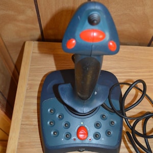 May include: A dark blue joystick with a black handle and red buttons. The joystick has a base with several buttons and a central red button. A black cable extends from the base.