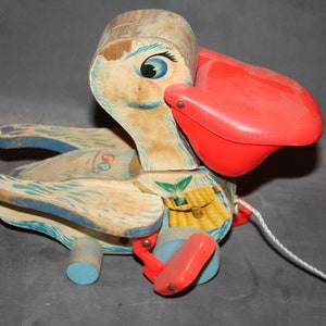 May include: Vintage wooden pull-along toy in the shape of a pelican. The toy features a red beak, blue and white painted details, and a white pull string. The toy has a basket design on its body and blue wheels.