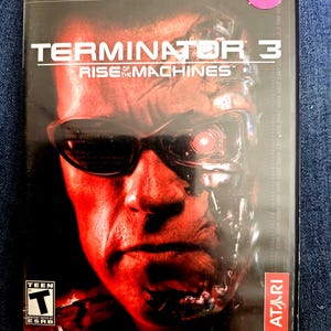 May include: A PlayStation 2 video game case for Terminator 3: Rise of the Machines. The cover features a close-up of a face with red lighting, wearing dark sunglasses with a glowing red eye visible. It includes the ESRB Teen rating and Atari logo.